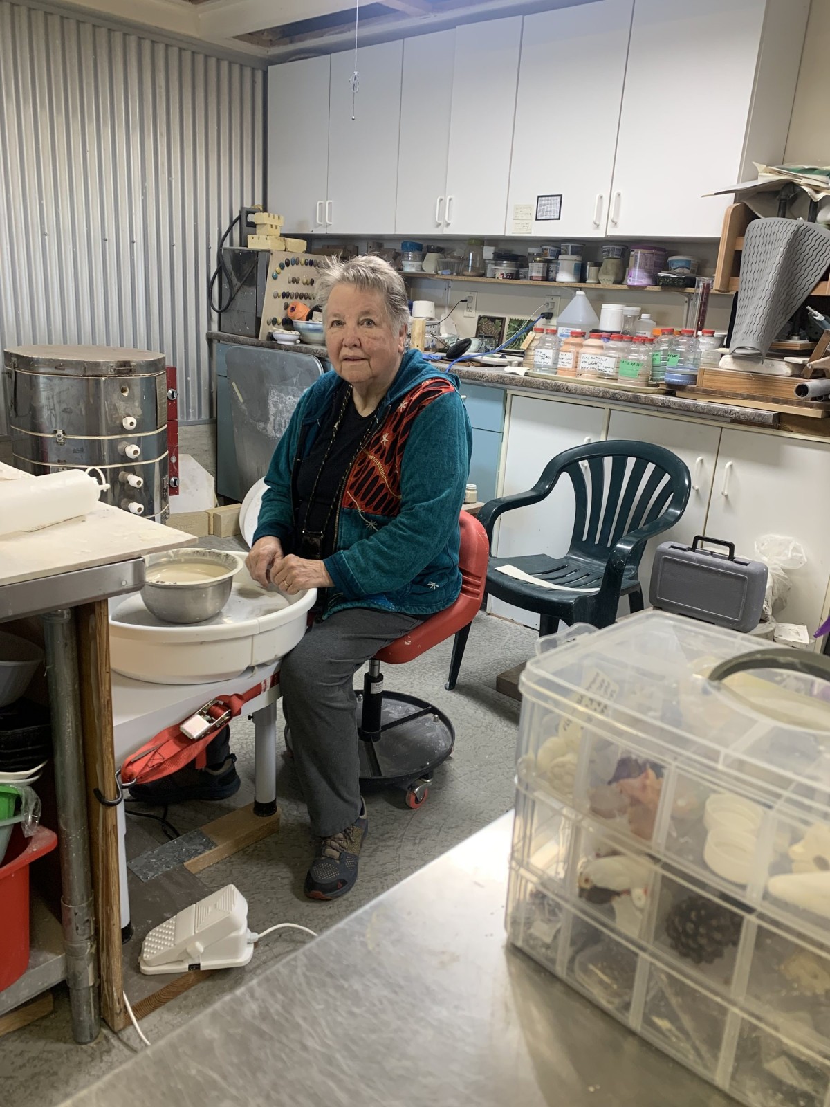 Kathleen Blohm seated at her work table in the studio, glaze jars and tools on the shelves behind her