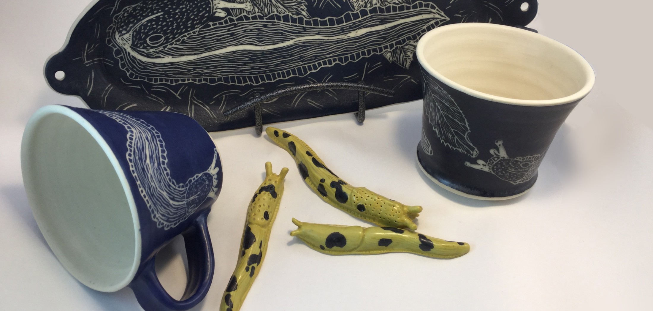 A hand-built platter, mug and beaker in dark blue sgraffito with banana slug drawings, and three small yellow clay slugs, on a white background