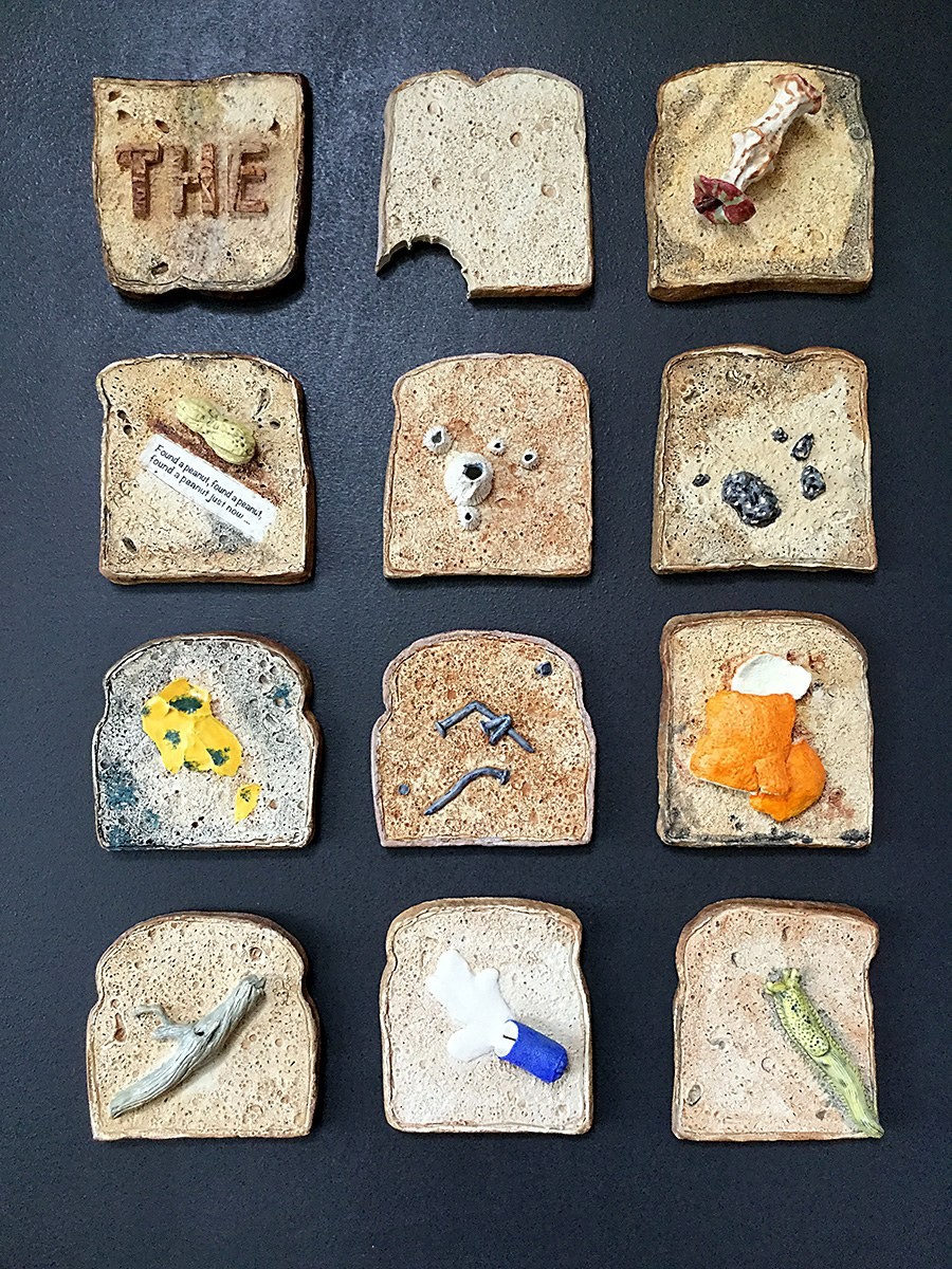 Twelve clay slices of toast on a dark ground, each carrying a different small object or spill — a peanut, a bone, jam, an egg, a slug