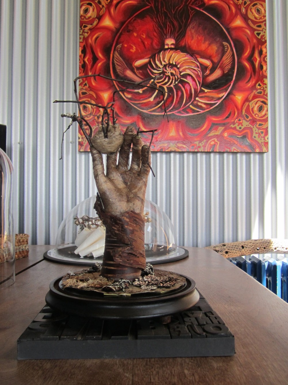 Sculpture of a weathered bark covered hand reaching upward from a bark base, holding a wasp nest and bare twigs, mounted on a dark bell jar base