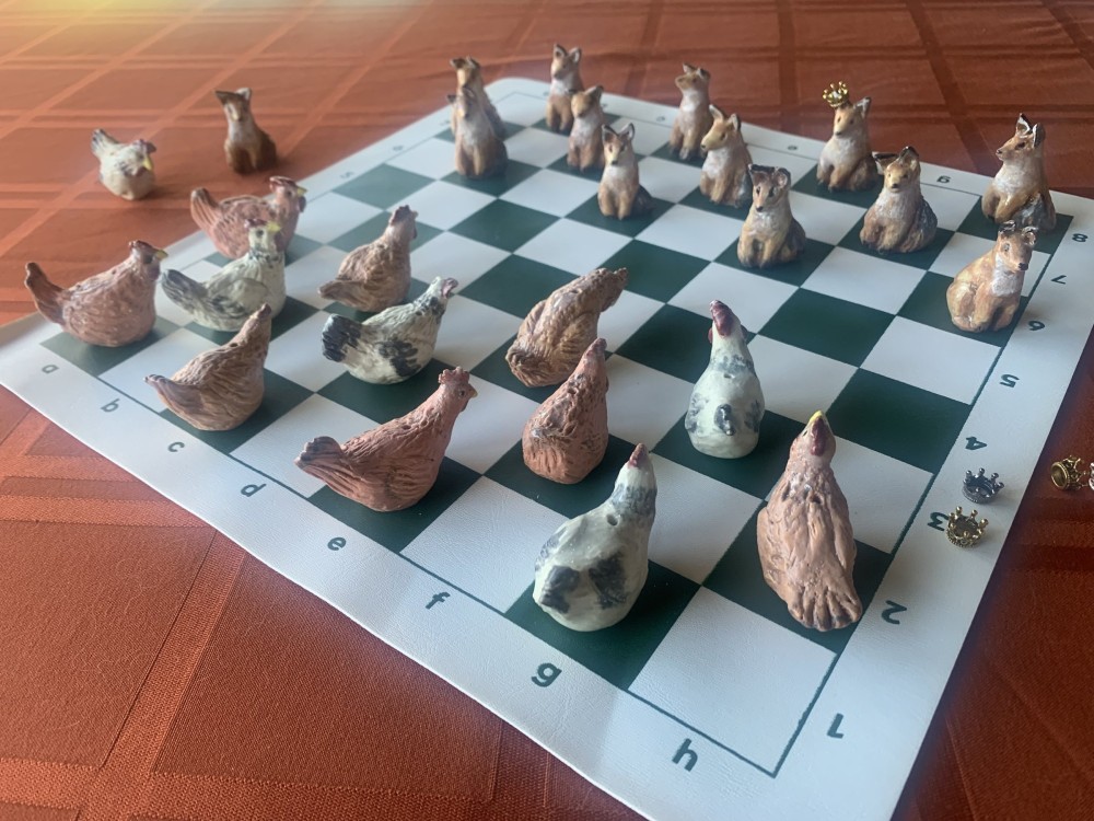 A checker set of small clay figures laid out on a board — hens on one side, foxes on the other, the kinged pieces wear tiny gold crowns