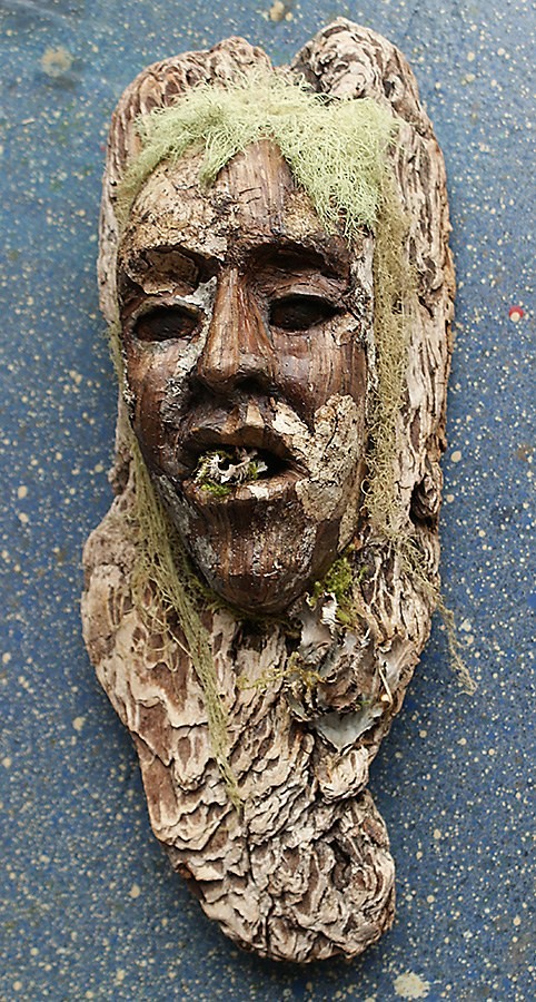 bark mask with a carved face, pale green lichen falling either side like long hair, and moss in the open mouth