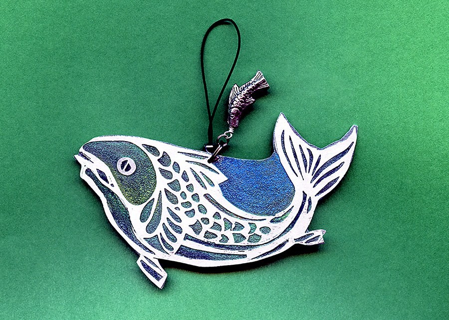 A single etched metal fish ornament with scales and fins etched through the surface, glazed blue and green, hanging from a cord with a small pewter fish charm
