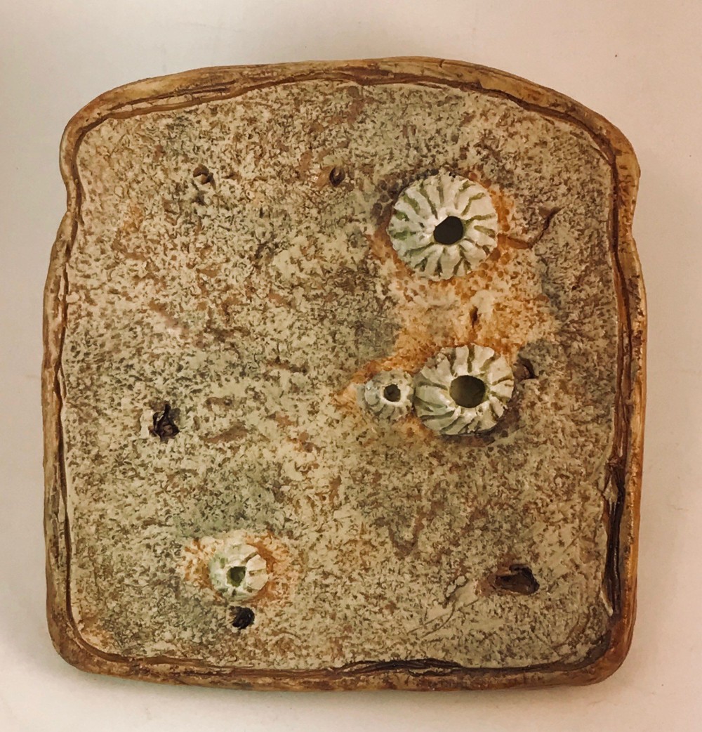 Stoneware slice of toast in speckled grey-green with three white barnacles growing on its surface