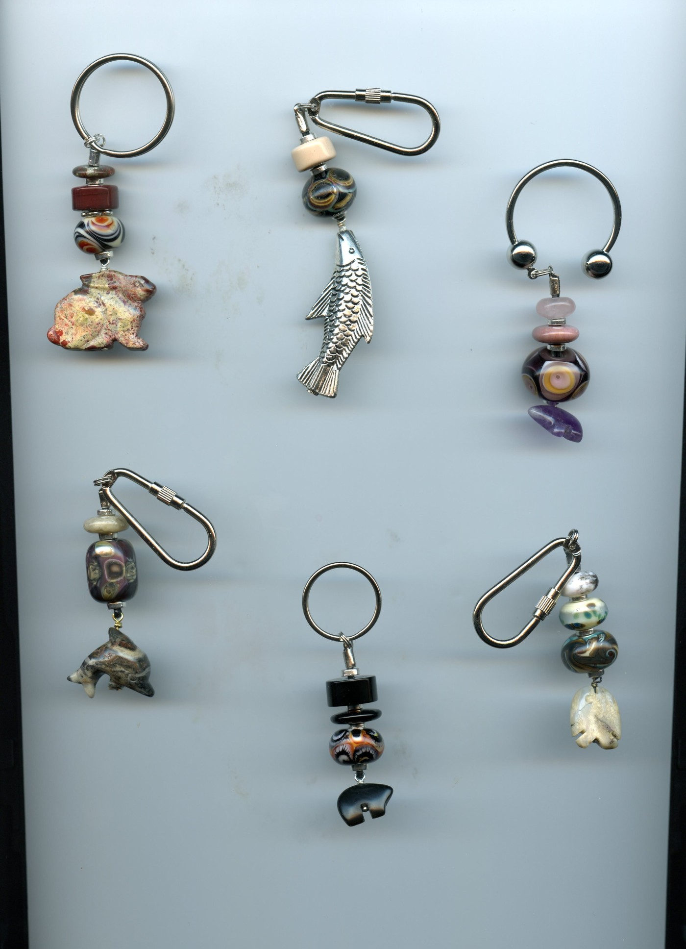 Six keychains laid out on a light ground, each combining lampwork glass beads with a carved stone or pewter charm — a bear, a dolphin, a fish, a rabbit
