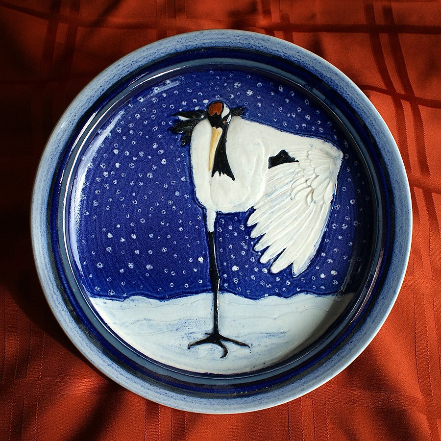 Round plate glazed blue and white, a crane with a raised wing standing in snow beneath falling snowflakes